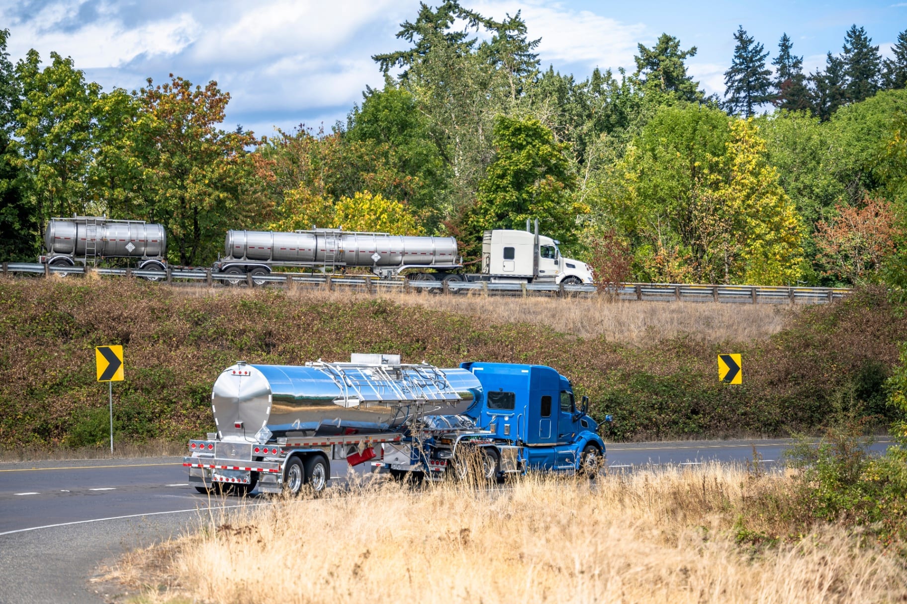 Blue tractor with stainless bulk tank trailer on a tree-lined highway