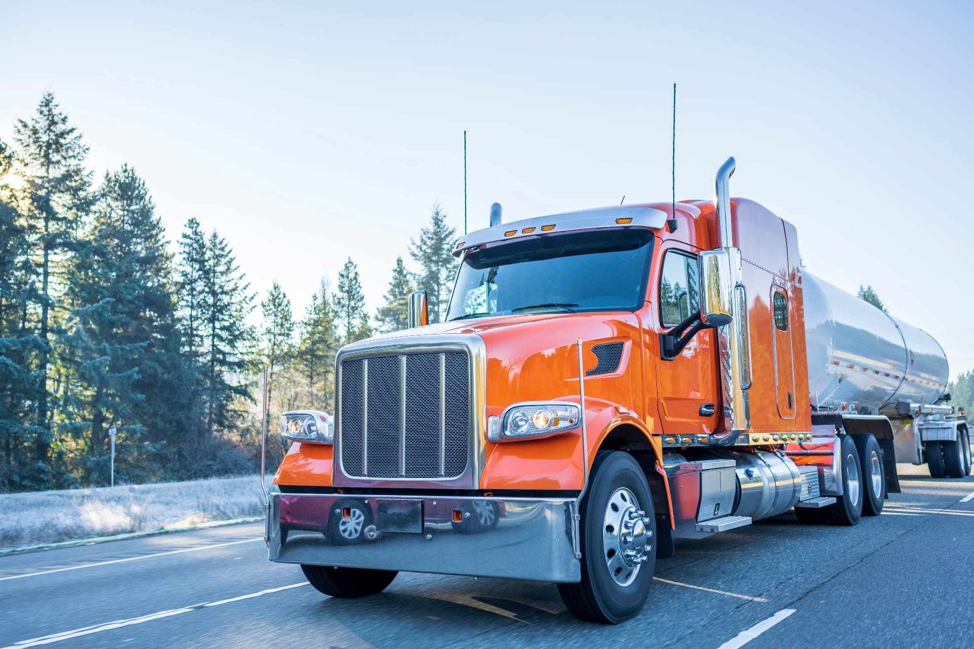 Orange tractor pulling a stainless bulk tank trailer on a forested highway